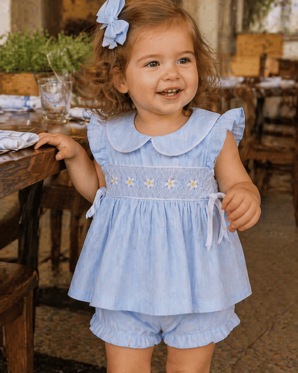 Child wearing a light blue dress with floral embroidery in an outdoor setting.