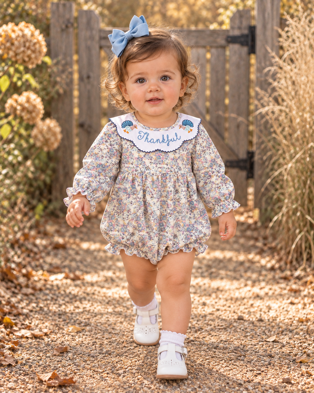 A toddler girl walking on a gravel path outdoors, wearing a long-sleeve floral romper with a white collar embroidered with the word 'Thankful' and blue birds, a light blue bow in her hair, and white shoes.