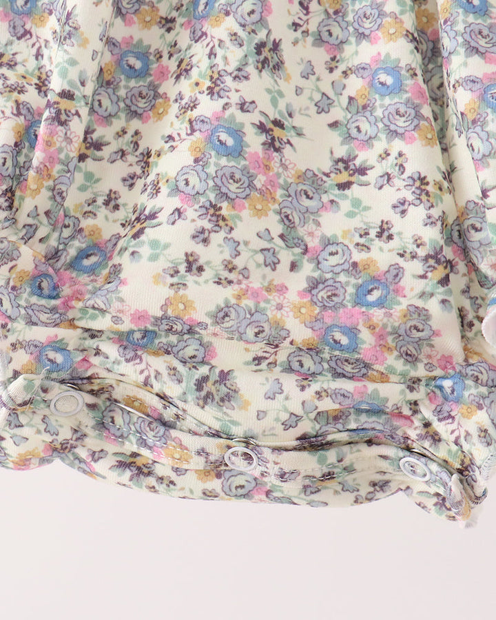 Close-up of a floral-patterned fabric with a ruffled hem, featuring a delicate print of blue, pink, yellow, and purple flowers on a cream background.