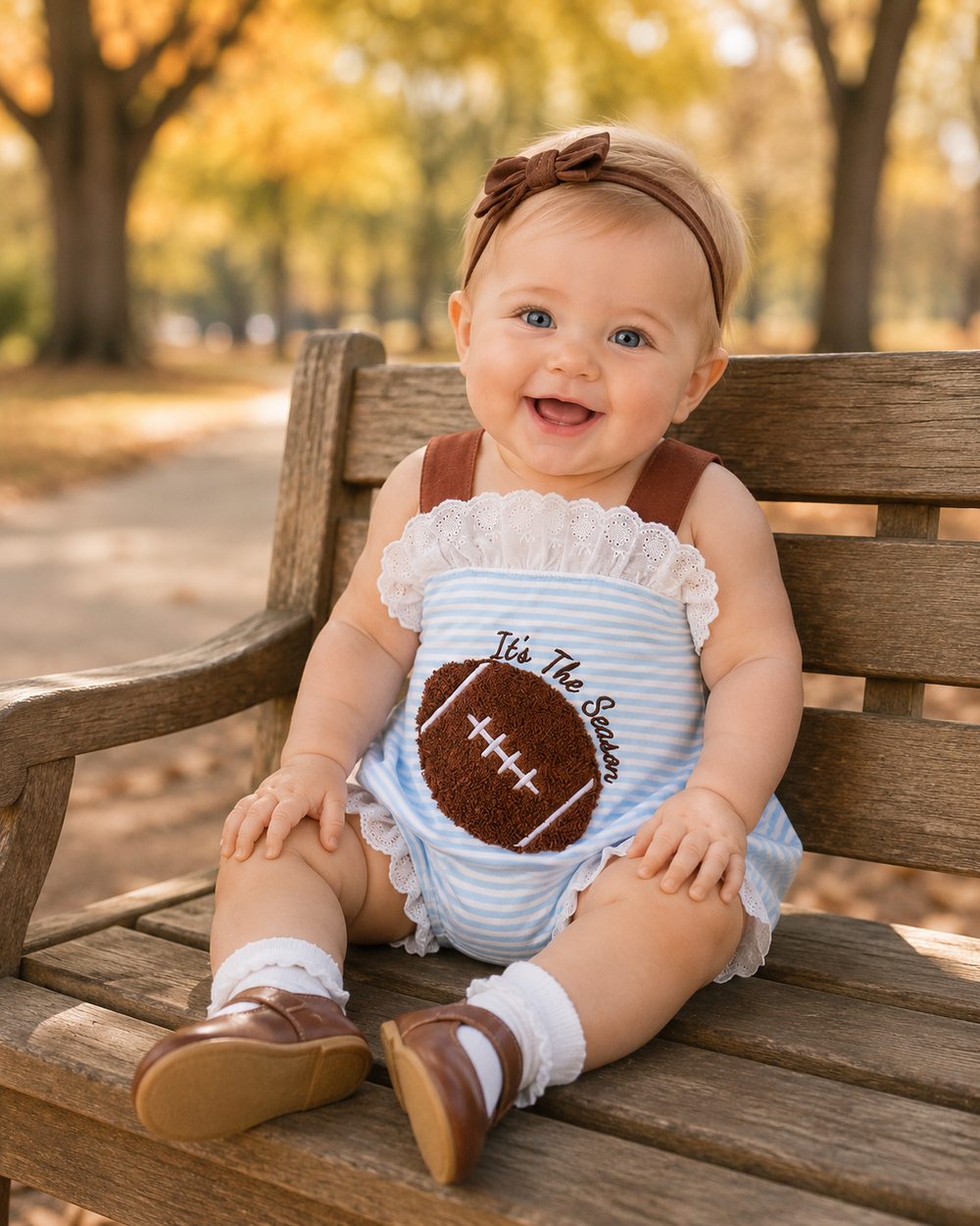 A baby sitting on a wooden park bench wearing a light blue and white striped romper with a brown football applique and the text 'It's The Season', along with a brown headband and brown shoes.