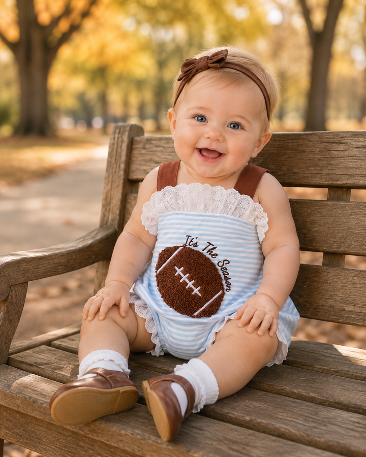 A baby sitting on a wooden park bench wearing a light blue and white striped romper with a brown football applique and the text &