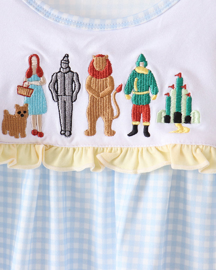 A close-up of a white baby top or nightgown featuring embroidered characters from The Wizard of Oz across the chest, finished with a pale yellow ruffle and a light blue and white gingham patterned hem.