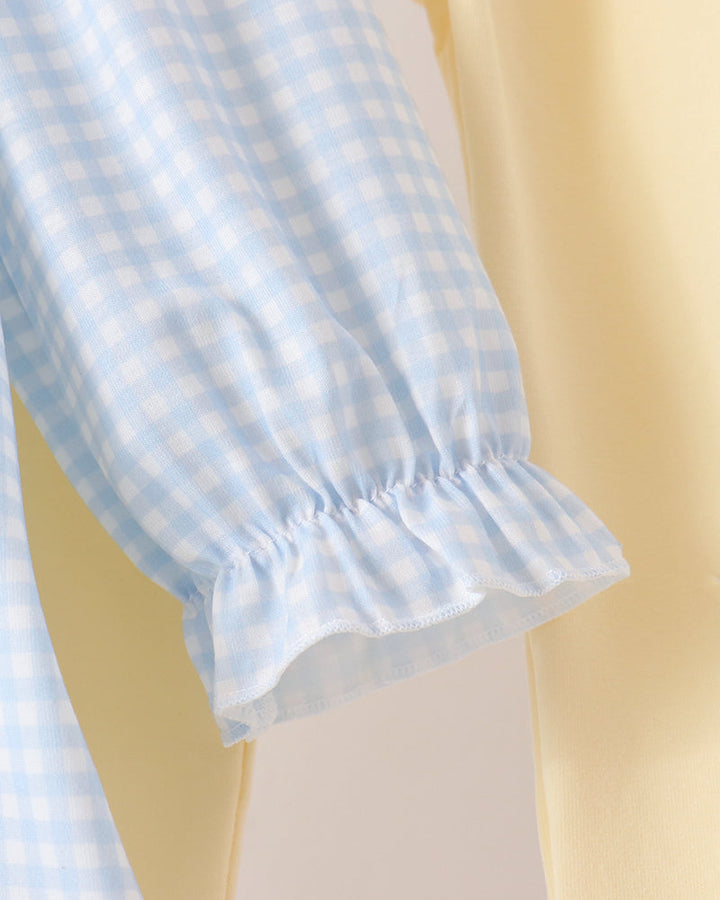Close-up of a light blue and white gingham checkered fabric sleeve with a ruffled hem, layered over a soft pale yellow garment.