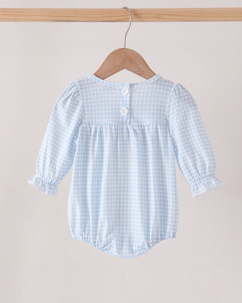 A baby romper in a light blue and white gingham pattern, hanging on a wooden hanger from a wooden clothes rack against a solid light beige background.