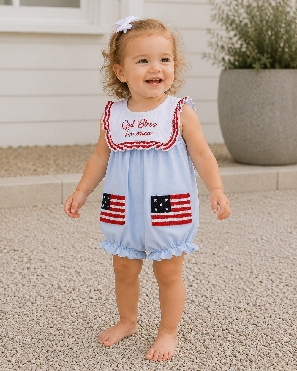 Child wearing a light blue romper with American flag designs and 'God Bless America' text outdoors.