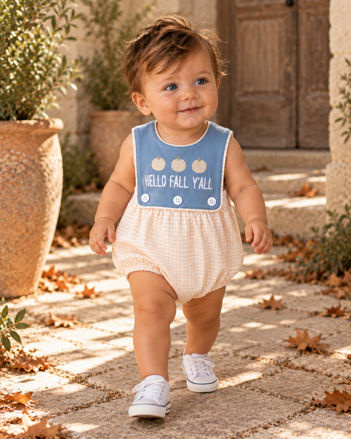 A toddler walking outdoors on a sunlit stone path wearing a sleeveless romper with a light blue bib-style front panel that says “HELLO FALL Y’ALL” and a cream gingham body.