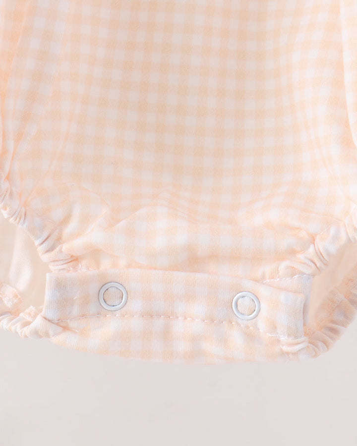 Close-up of a baby romper or bodysuit fabric with a pale peach and white gingham check pattern, featuring elasticized ruffled edges and two white snap closures at the crotch.