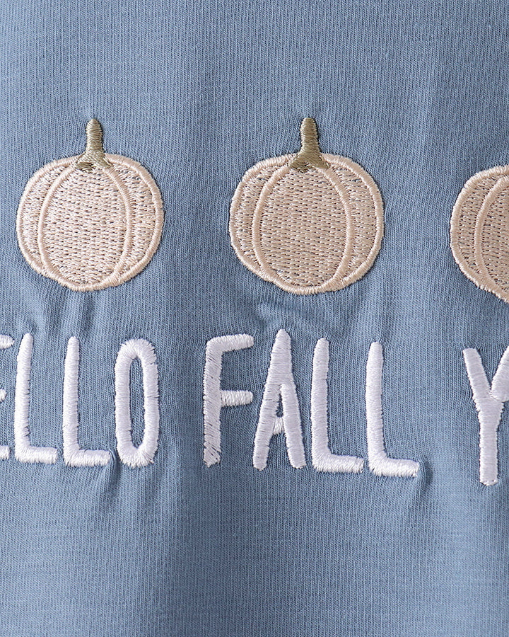 Close-up of a light blue fabric garment with embroidered cream pumpkins and white text reading &