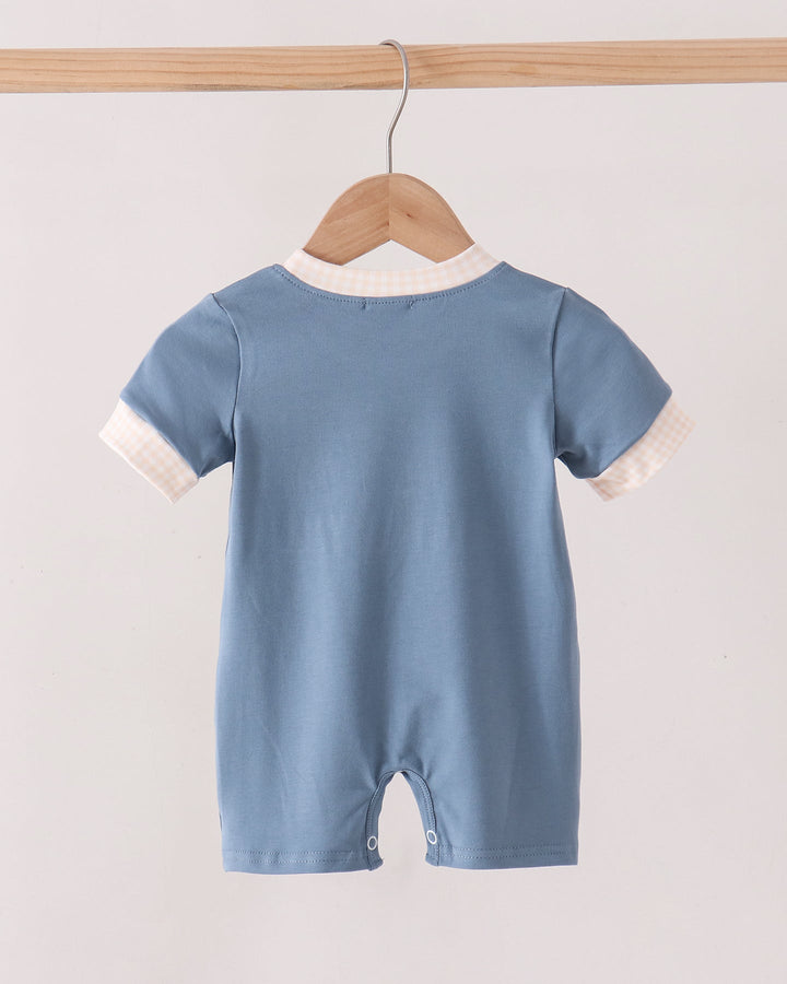 A light blue baby romper with white contrast trim on the collar and sleeve cuffs, hanging on a wooden hanger from a wooden clothes rack against a plain off-white background.