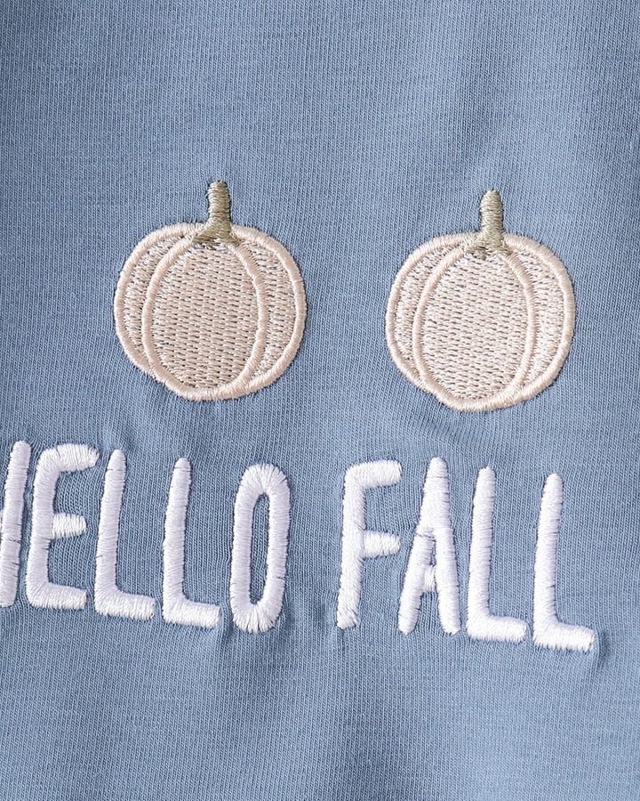 Close-up of a light blue fabric surface with two embroidered cream pumpkins above white embroidered text reading &
