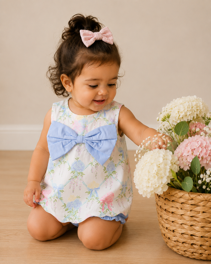 Child in a floral dress with a large bow sitting next to a basket of flowers on a wooden floor.