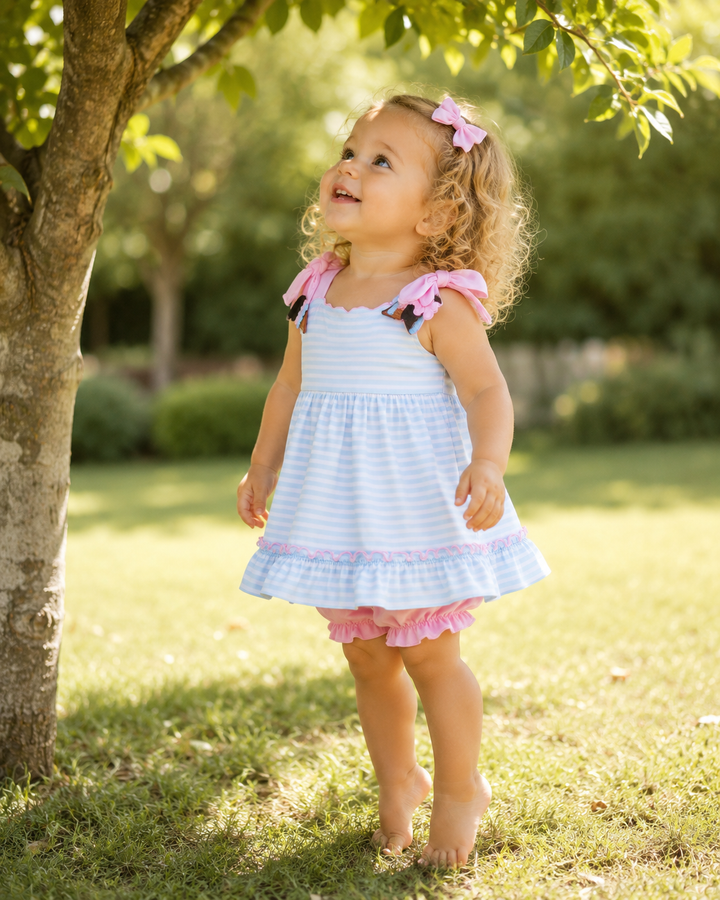 Young girl in a light blue dress with pink accents standing under a tree in a park.