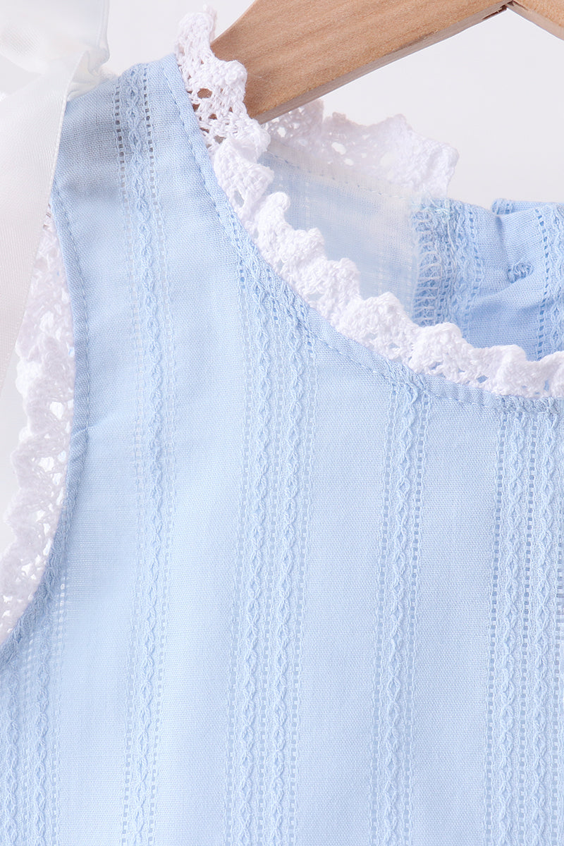 A close-up of a light blue sleeveless top with vertical eyelet embroidery and white lace trim, hanging on a wooden hanger against a white background.