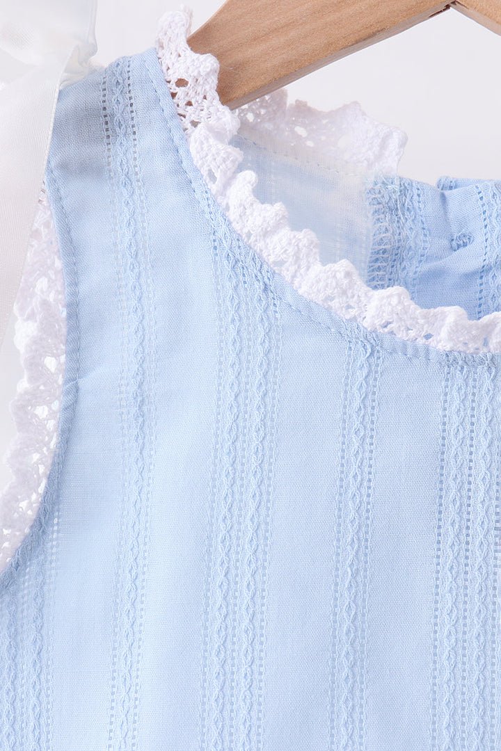 A close-up of a light blue sleeveless top with vertical eyelet embroidery and white lace trim, hanging on a wooden hanger against a white background.