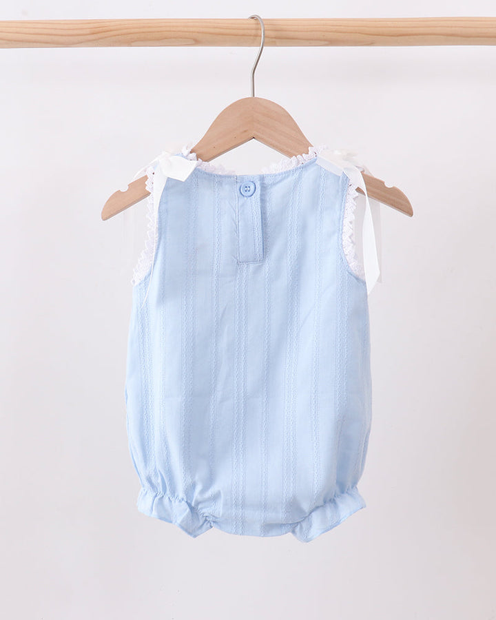 A light blue sleeveless baby romper with white lace shoulder details, hanging on a wooden hanger from a wooden clothes rack against a plain off-white wall.