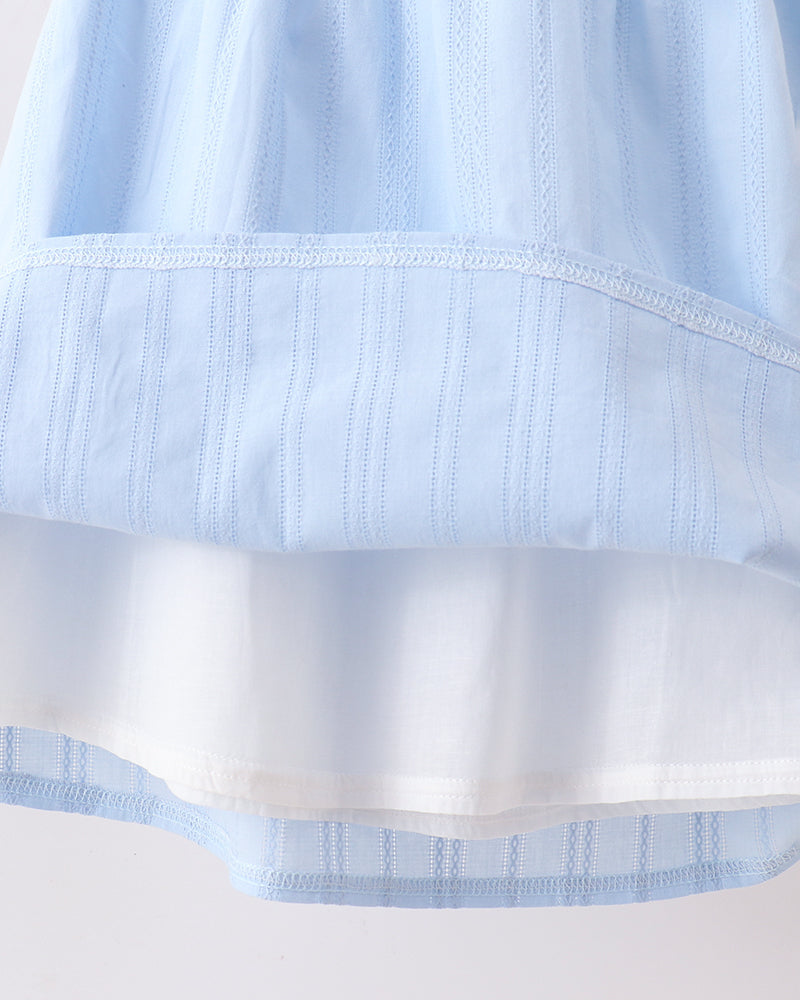 Close-up of a light blue and white tiered fabric garment, showing the layered construction and fine vertical embroidery.
