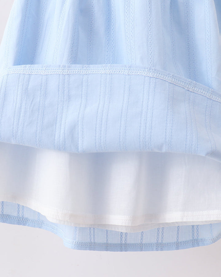 Close-up of a light blue and white tiered fabric garment, showing the layered construction and fine vertical embroidery.
