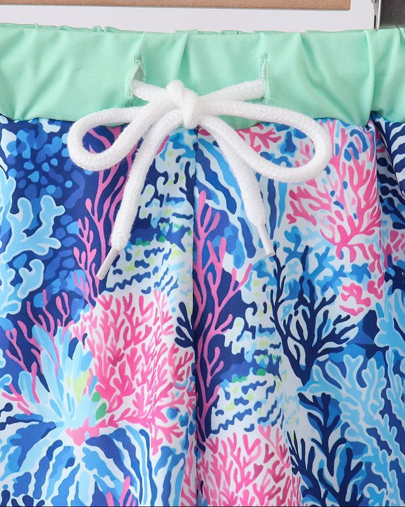 Colorful swim trunks with a coral pattern on a wooden hanger against a white background