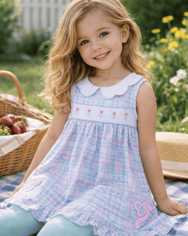 Young girl in a plaid dress sitting outdoors with a picnic basket and flowers.