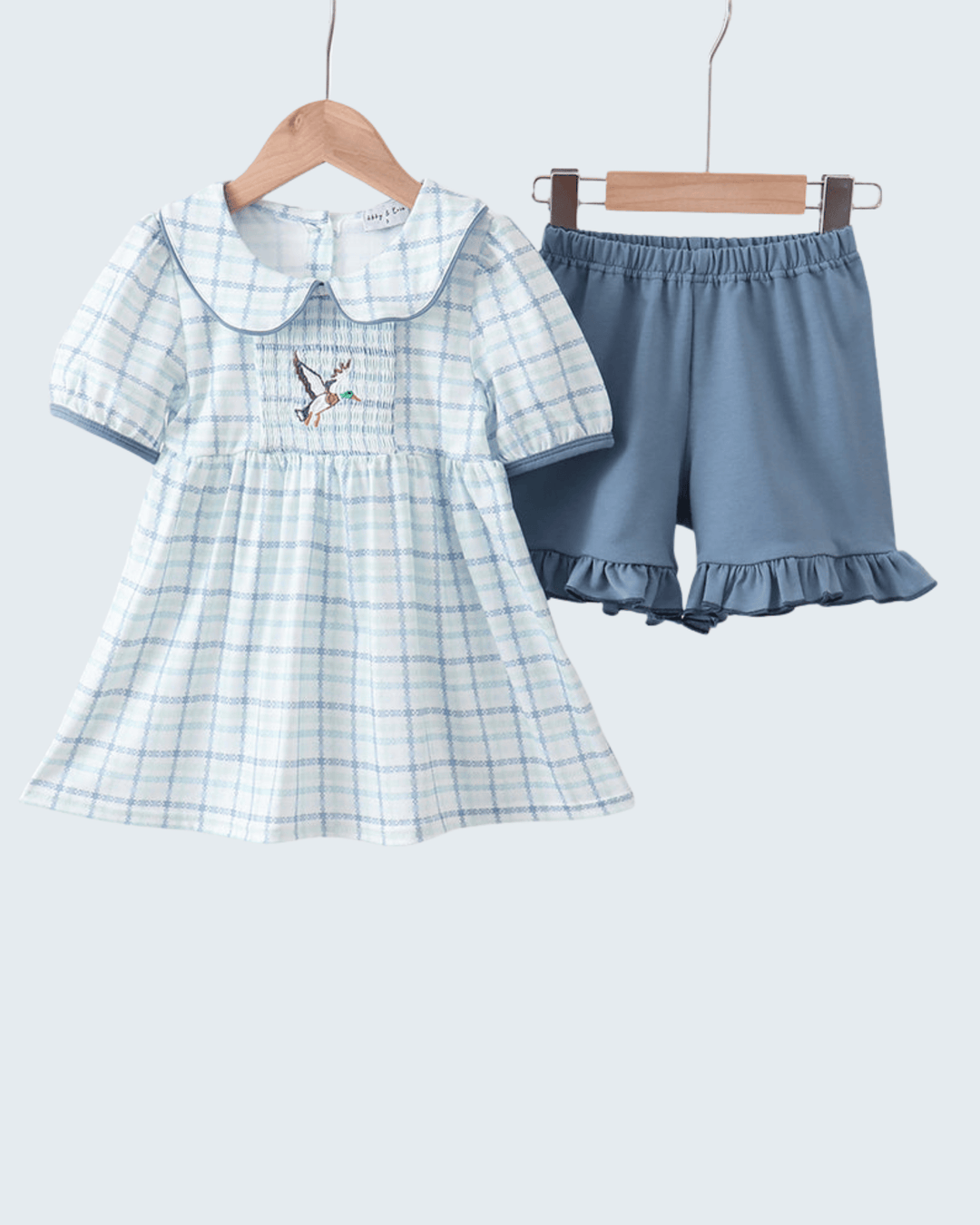 Children's blue plaid dress with embroidered design and matching shorts on hangers against a white background