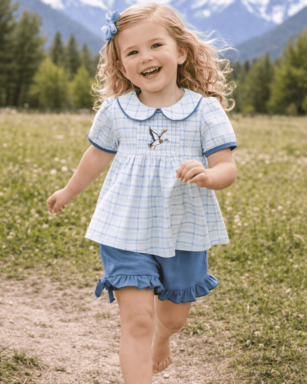 Child in a blue checkered dress with a bird design, standing in a field with mountains in the background.