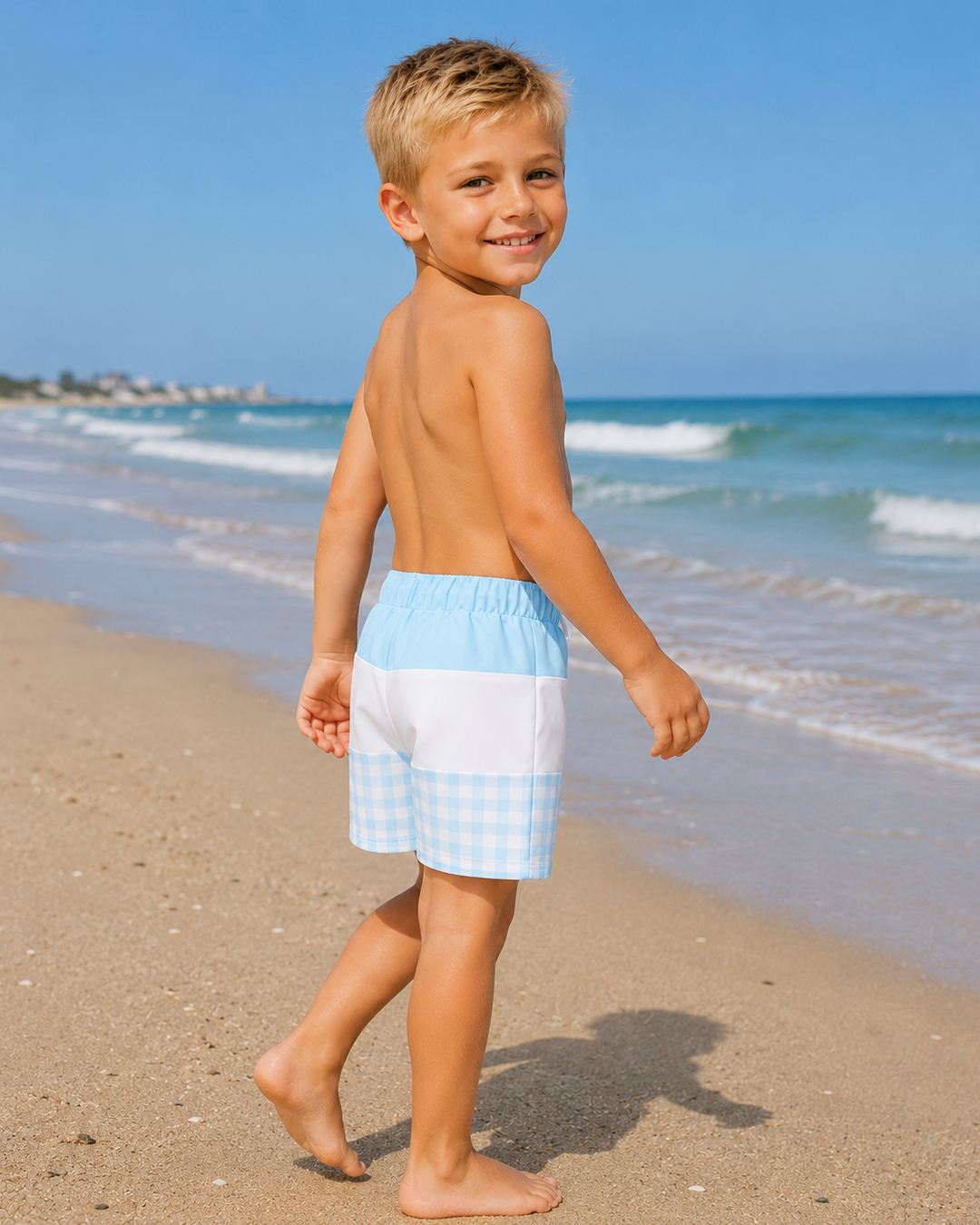 Young boy in checkered swim shorts walking on a beach with ocean waves in the background