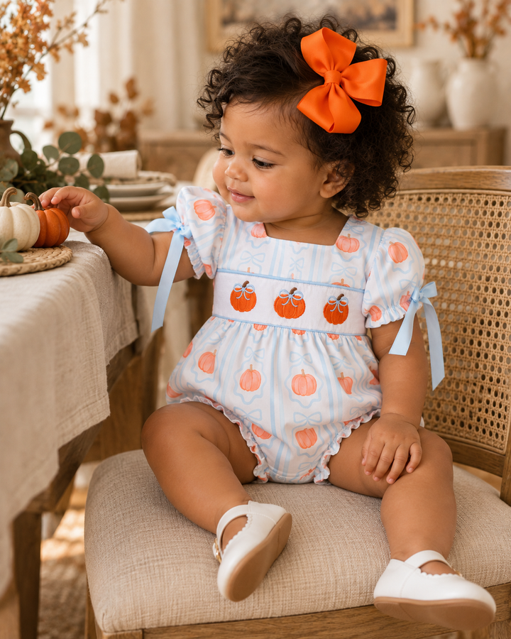 A toddler girl with curly dark hair wearing a large orange bow sits on a woven chair in a warm, decorated indoor setting. She is wearing a light blue and white patterned romper with orange pumpkins and blue ribbon ties, along with white shoes.