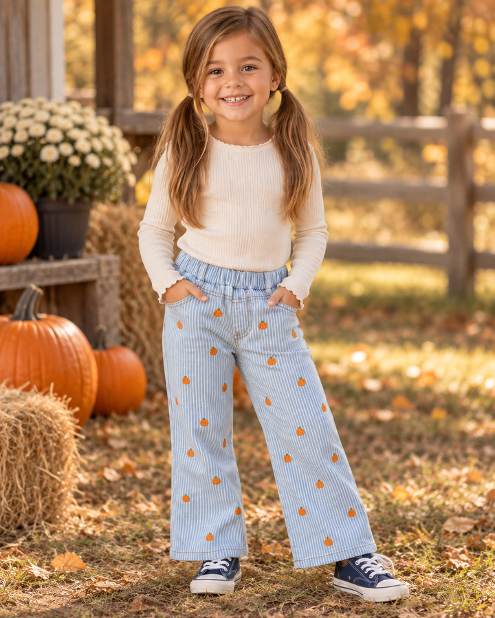 A young girl with pigtails stands outdoors in a fall yard wearing a cream ribbed long-sleeve top and light blue pinstriped wide-leg pants with orange pumpkin prints. She is standing on grass with pumpkins, hay bales, and a wooden fence in the background.