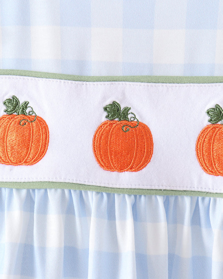 Close-up of a baby dress featuring a light blue and white gingham pattern, a white smocked band with embroidered orange pumpkins and green stems, and a ruffled hem.
