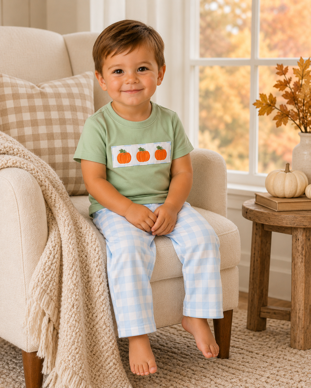 A young child sitting on a beige armchair in a cozy room, wearing a light green t-shirt with three orange pumpkins on a white rectangular patch and light blue and white gingham pants.
