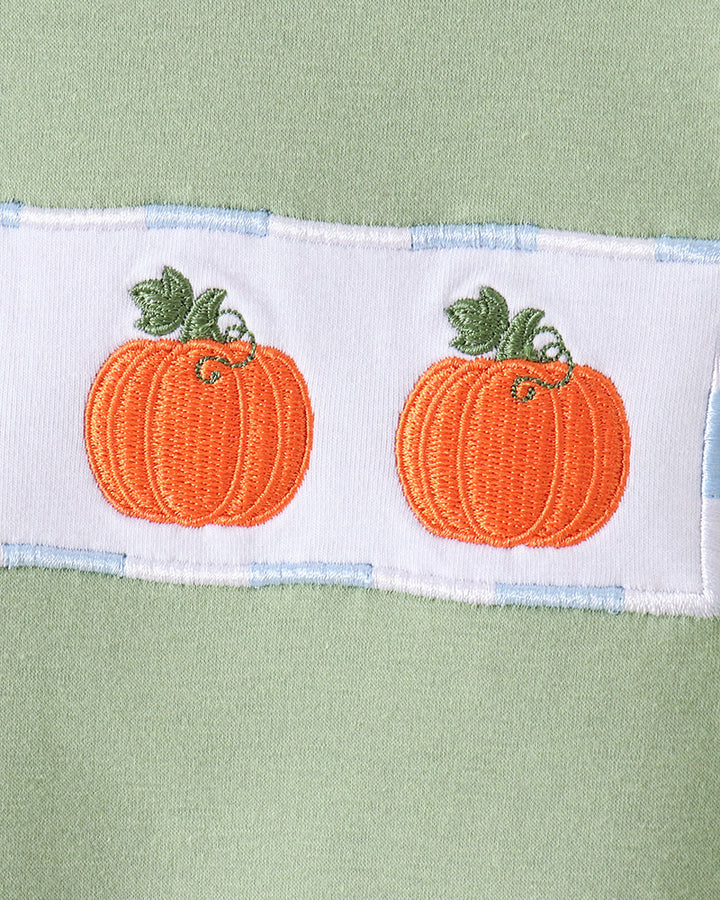 A close-up of a light sage green fabric garment featuring a white horizontal band with two embroidered orange pumpkins and green stems.