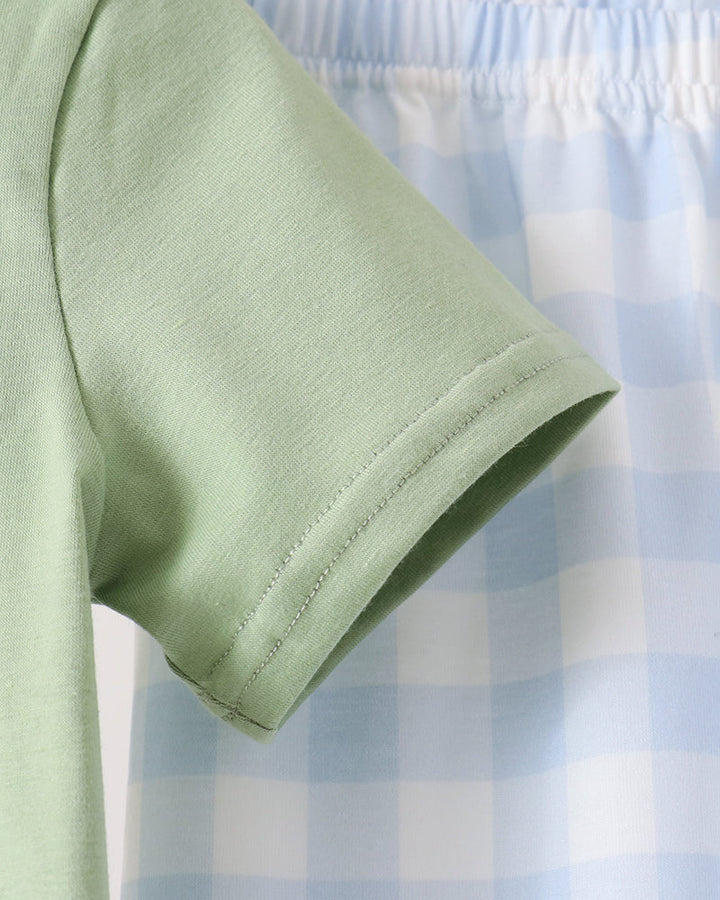 Close-up of a light sage green short-sleeve t-shirt hanging on a wooden hanger, with a light blue and white gingham patterned garment partially visible behind it.