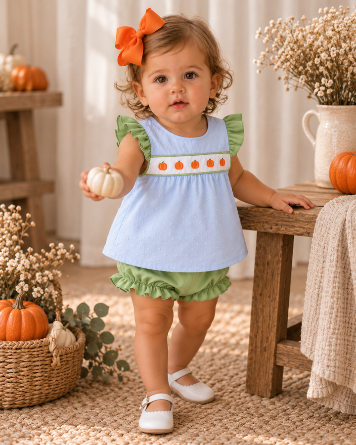 A toddler stands indoors wearing a light blue sleeveless top with green ruffle trim and a white chest band decorated with orange pumpkin motifs, paired with green ruffled bloomers and white shoes. The child is posed beside a wooden table with pumpkins, dried flowers, and a woven rug in a warm autumn setting.