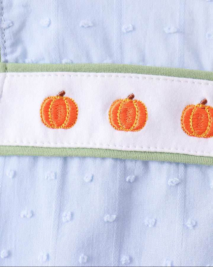 Close-up of a baby garment featuring a light blue textured fabric, a white horizontal band with three orange pumpkin motifs, and a thin sage green trim.
