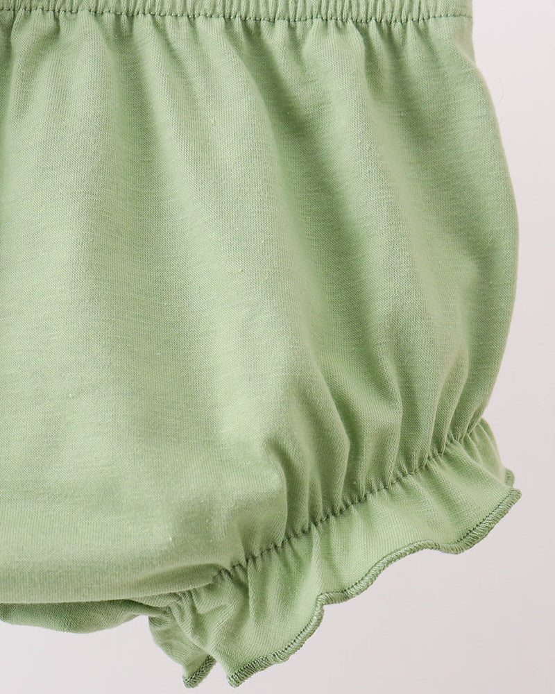 A close-up of light sage green fabric, likely a baby bloomers or diaper cover, showing the elastic waistband and scalloped ruffle hem against a plain light background.
