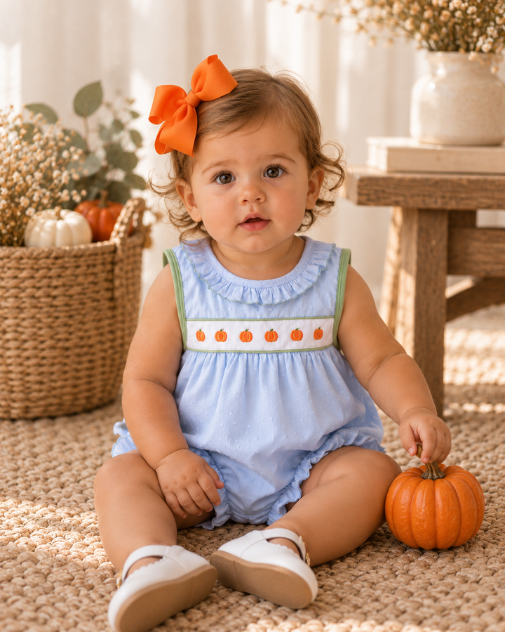 A baby girl sitting on a beige rug wearing a light blue romper with a white smocked chest panel decorated with orange pumpkins, a green shoulder strap, and an orange bow in her hair. She is holding a small orange pumpkin next to a wicker basket with pumpkins.