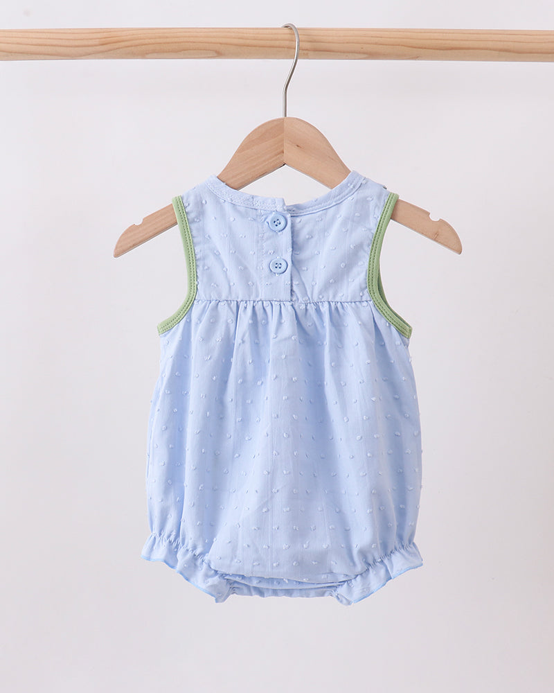 A light blue baby romper with tiny white polka dots, hanging on a wooden hanger from a wooden clothes rack. The romper features green trim along the shoulder seams and a button closure at the back.