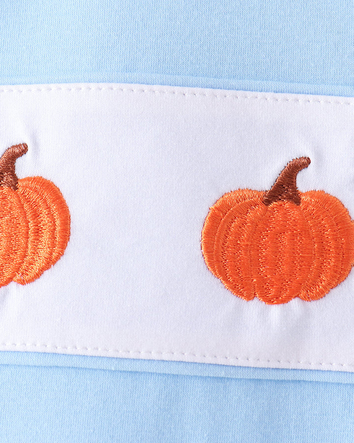 Close-up of a light blue garment with a white horizontal band featuring embroidered orange pumpkins with brown stems.