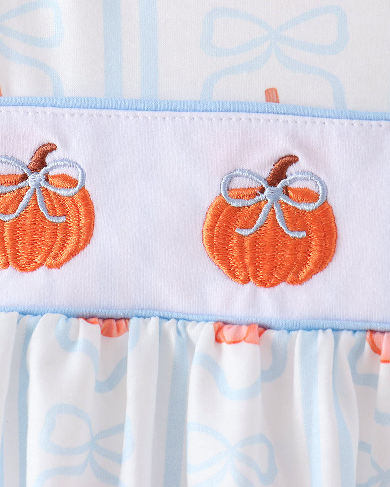 Close-up of a baby garment featuring orange pumpkin embroidery with light blue bows on a white band, with a white skirt printed with light blue bows and orange pumpkins.