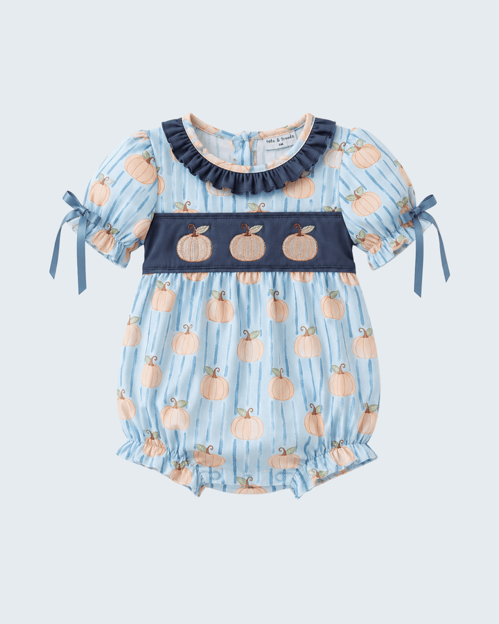 A baby romper in light blue with vertical stripes, featuring a pumpkin print, a navy blue smocked chest panel with three embroidered pumpkins, and navy blue ruffled collar. The short sleeves have navy blue ribbon ties on the sides.