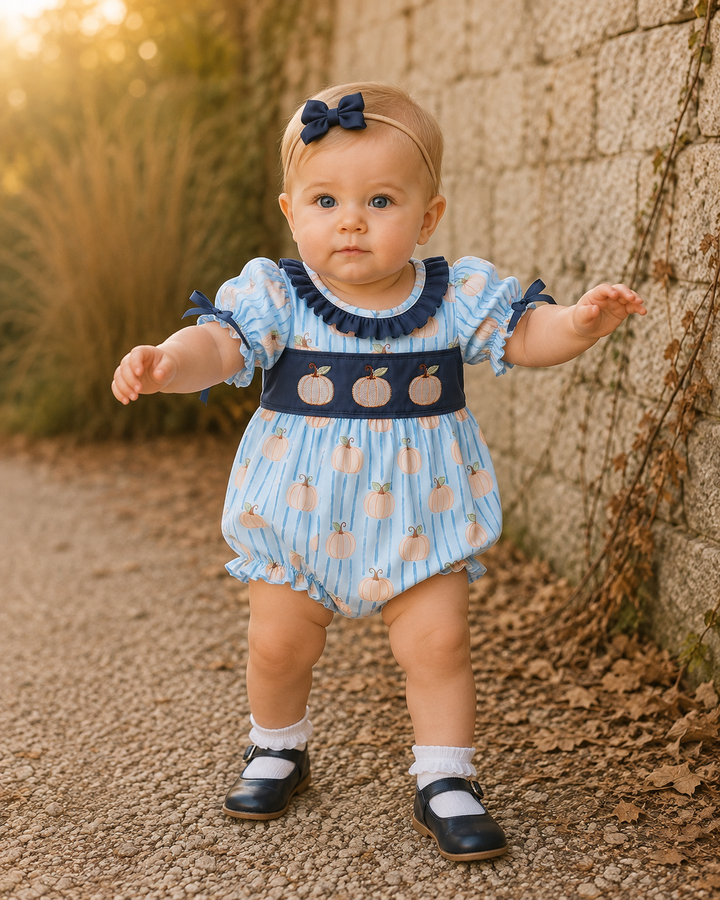 A baby girl wearing a light blue and navy blue romper with a peach pattern, a matching navy bow headband, white socks, and navy shoes, standing on a gravel path next to a stone wall.