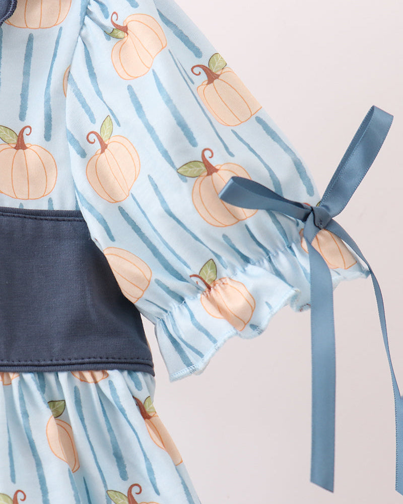 A close-up of a baby dress featuring a light blue and navy striped pattern with orange pumpkin prints, a solid navy waistband, and a matching navy ribbon bow on the sleeve.