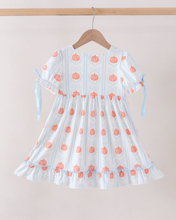 A short-sleeve dress with a white base, light blue vertical stripes, and a repeating orange pumpkin print, hanging on a wooden hanger against a light beige wall.