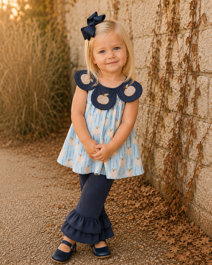 A young girl stands outdoors against a stone wall and dry plants, wearing a light blue and white patterned dress with navy ruffled collar details, navy ruffled pants, navy shoes, and a navy bow in her hair.