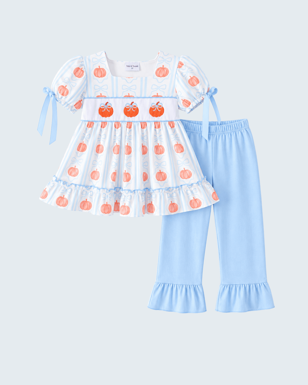 A toddler girl's two-piece clothing set featuring a white smocked dress with orange pumpkin prints and light blue trim, paired with light blue ruffled pants, shown on a light gray background.