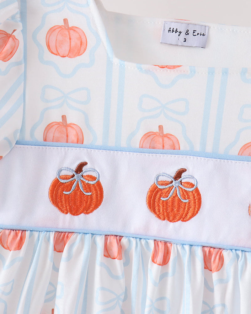 A close-up of a baby dress featuring a white base with light blue vertical stripes, orange pumpkin prints, and a smocked white band with two embroidered orange pumpkins wearing white bows. The brand label reads &