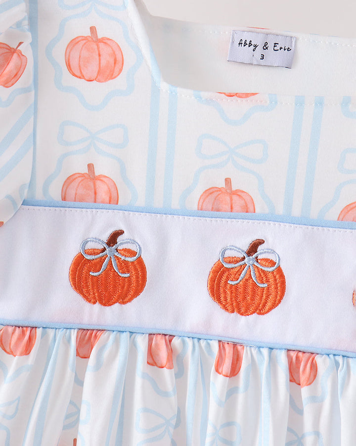 A close-up of a baby dress featuring a white base with light blue vertical stripes, orange pumpkin prints, and a smocked white band with two embroidered orange pumpkins wearing white bows. The brand label reads &