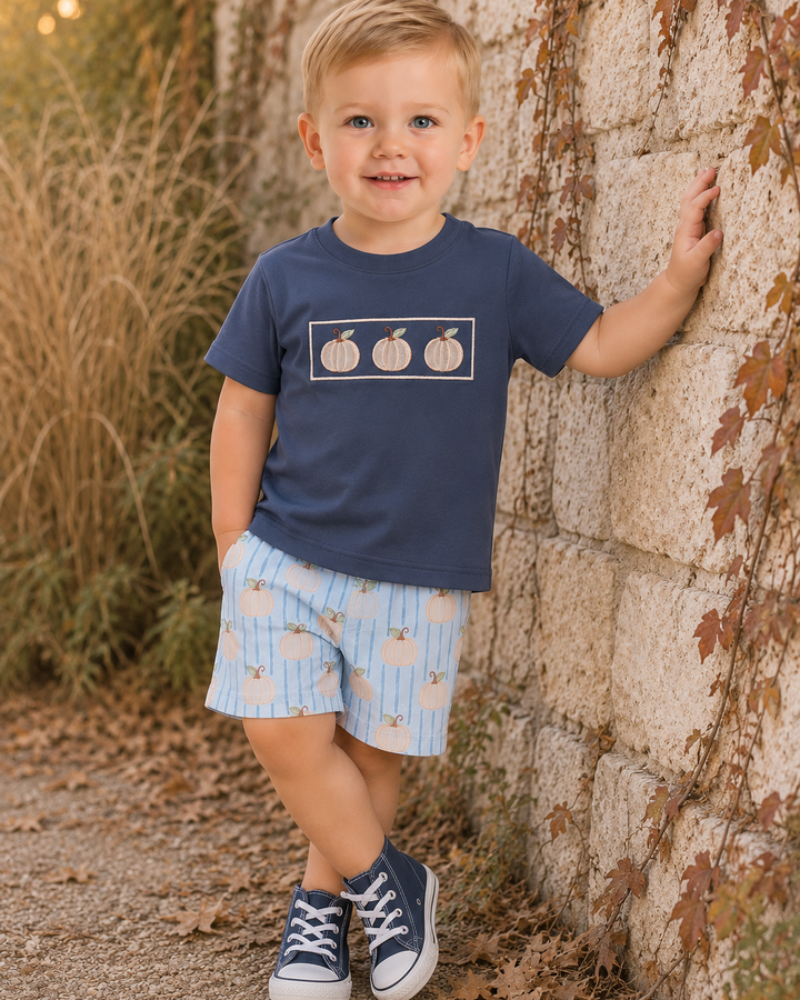 A young boy stands outdoors against a stone wall wearing a navy blue t-shirt with a pumpkin graphic and light blue patterned shorts.