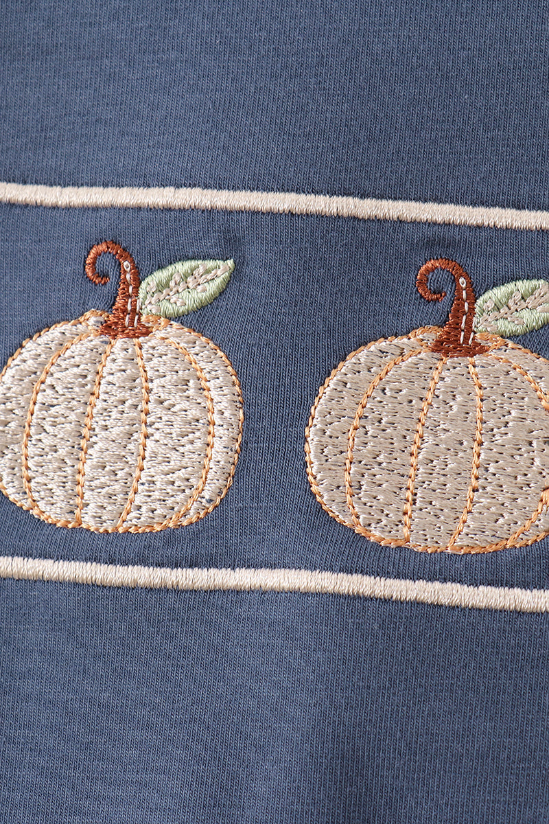 Close-up of a navy blue fabric garment featuring two embroidered cream pumpkins with brown stems and light green leaves, separated by thin cream horizontal stripes.