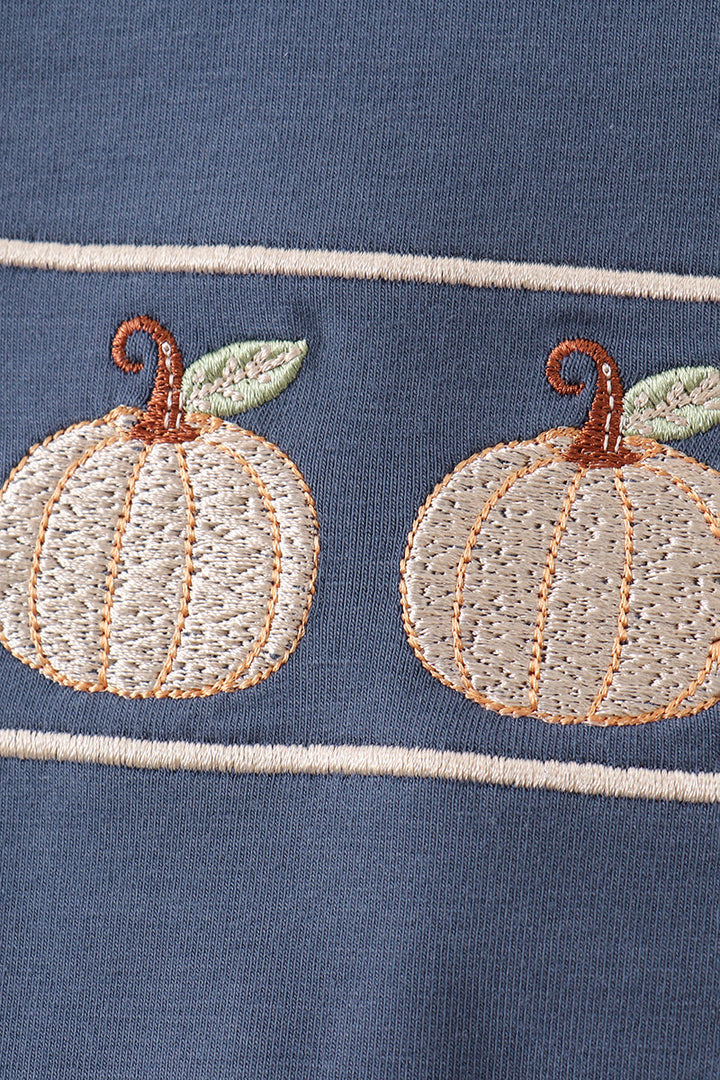 Close-up of a navy blue fabric garment featuring two embroidered cream pumpkins with brown stems and light green leaves, separated by thin cream horizontal stripes.